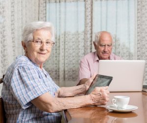 Older couple at a dining room table, using a laptop and a tablet. 